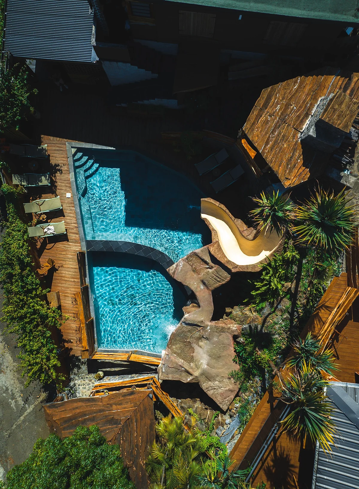 La piscine en rocaille vue du ciel, entre les palmiers et les terrasses en bois